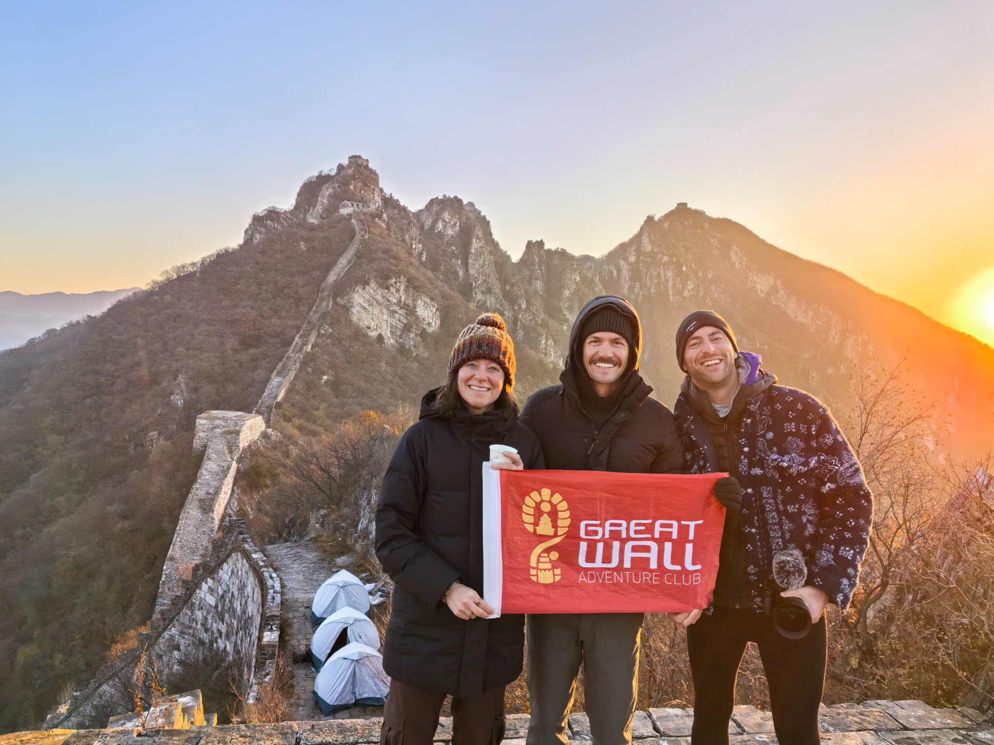 Three hikers holding a Great Wall Adventure Club flag beside the Great Wall at sunset