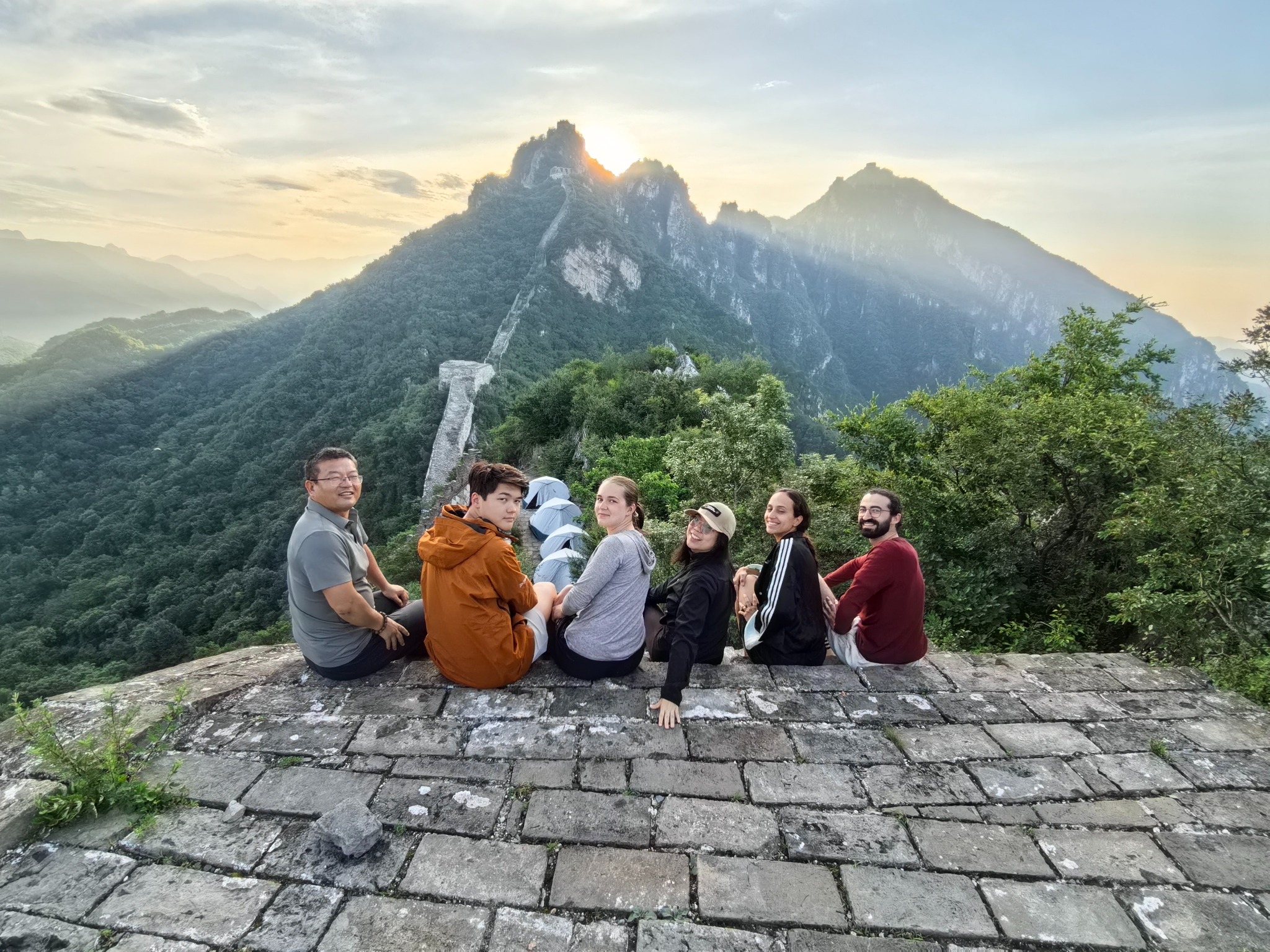 Six travelers sitting on the Great Wall with mountains and camping tents behind them